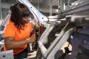 A young, Black female uses a hand-held tool to work on the metal interior frame of a car. She is wearing an orange, short-sleeve CU-ICAR polo shirt.