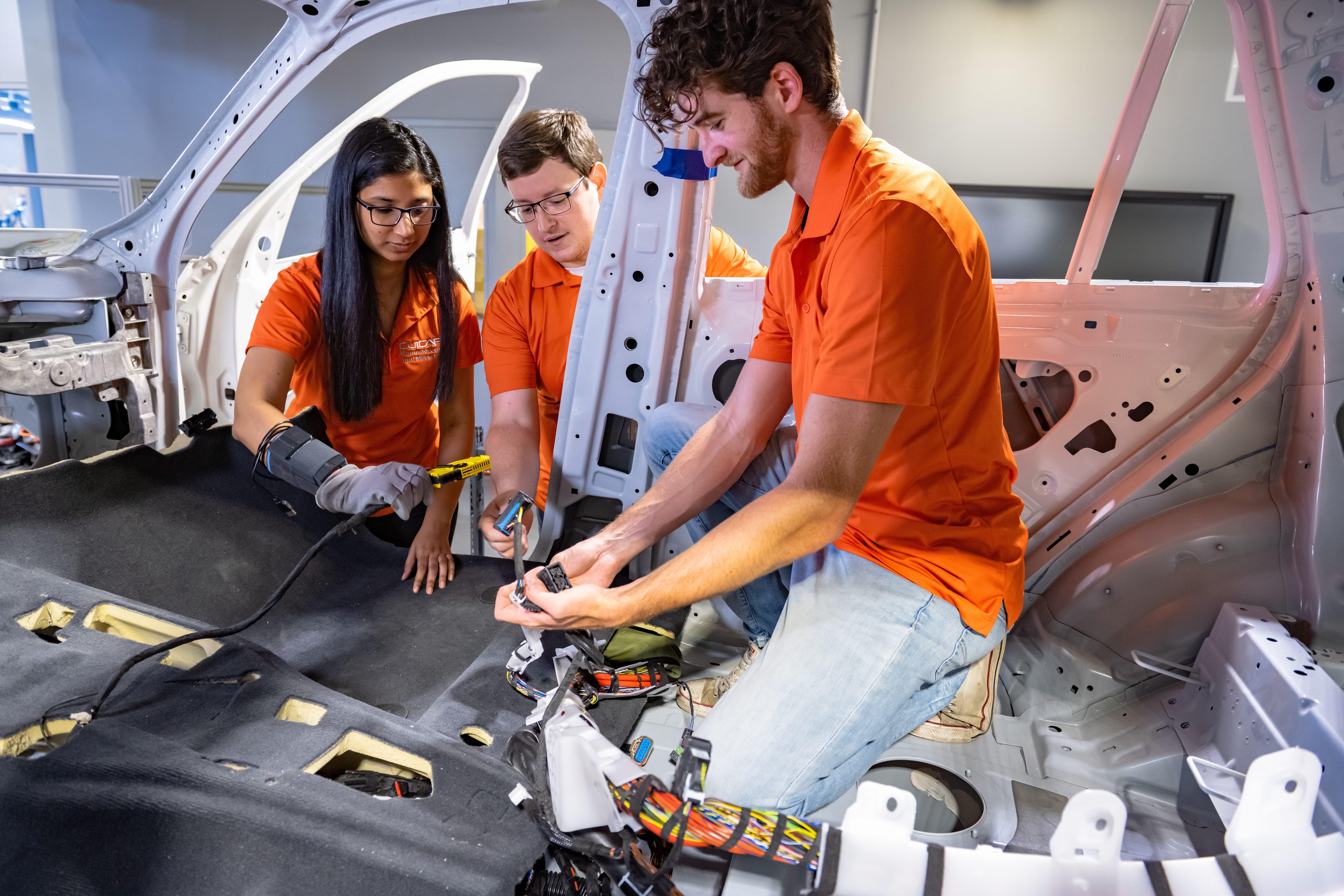 Two men and a women, all wearing orange, work on the inside of a vehicle's floorboard with hand-held tools.