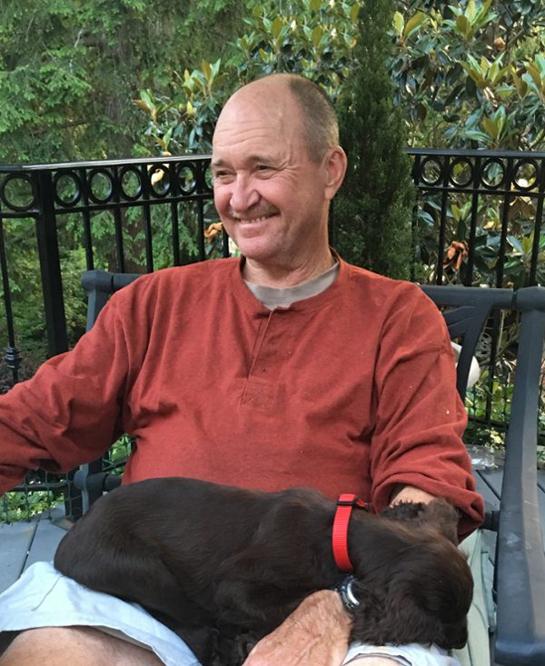 An older man, Mike Maloney, very short hair, nearly bald, and also a mustache, sits with a puppy in his lap. He is outside on a deck, wearing a smile and casual attire.