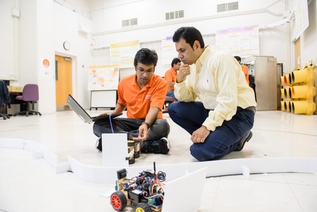 Two men, one a student, one a professor are in the middle of a large open space. The student is an Indian male wearing an orange, short-sleeve polo shirt and he is sitting cross-legged on the ground with a small, robotic vehicle in the foreground. The professor is crouched on one knee, wearing a long-sleeve yellow dress shirt and dark slacks. 