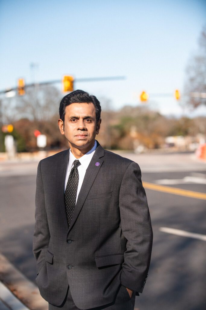 An Indian man in a dark suit and tie stands in the middle of a street with traffic lights behind him. 