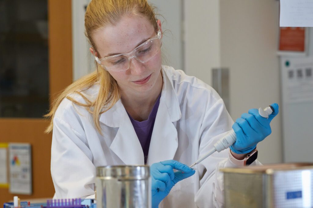 A young woman with a long, blonde ponytail wears a white coat, blue gloves and safety goggles, working in the lab.