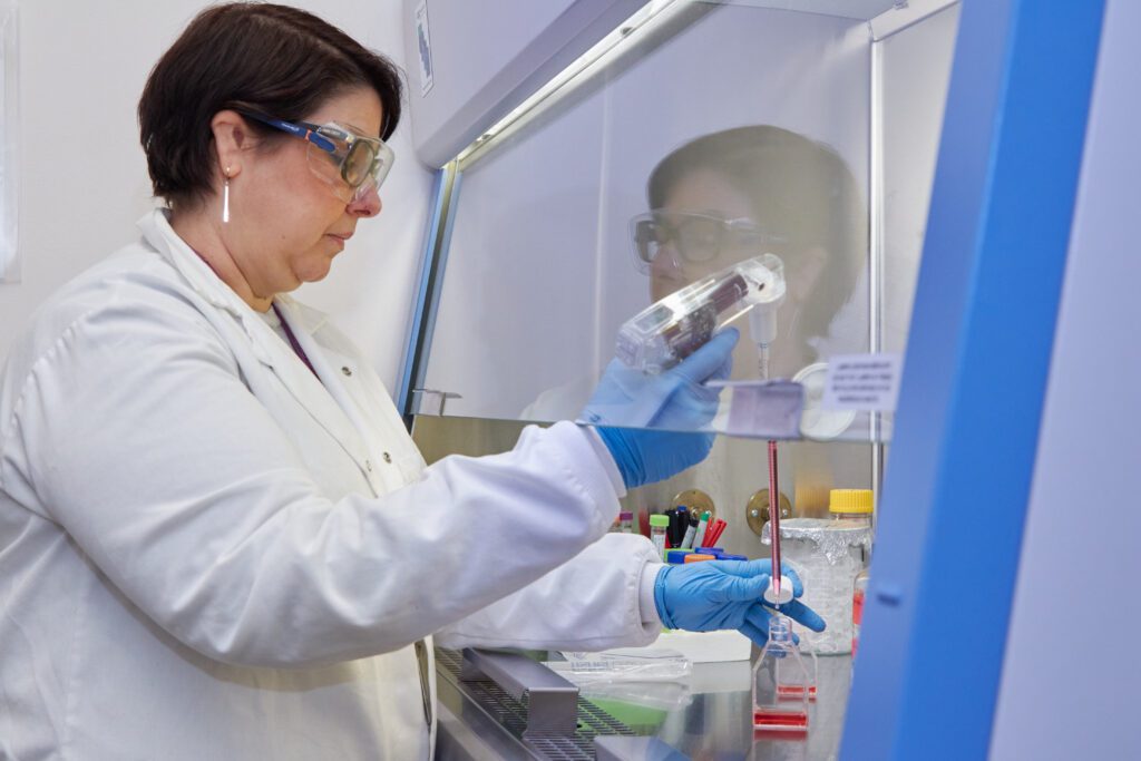 A female scientist wearing a white lab coat, safety goggles and blue gloves examines a tube from behind a clear divider.