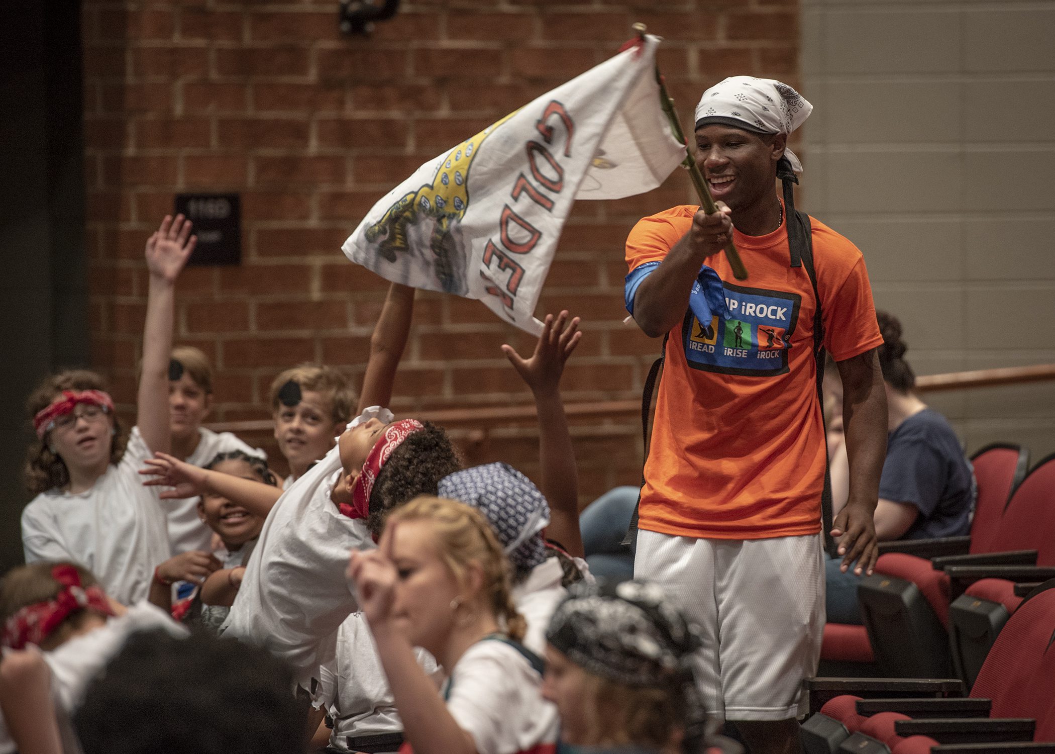 A young, Black male wearing an orange t-shirt waves a white flag that reads "Goldie" over a group of excited elementary-age children.