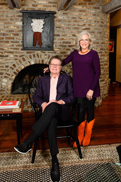 Thom and Gretchen Penney smile for a portrait. Thom is seated and Gretchen is standing. They are in front of a large brick fireplace.