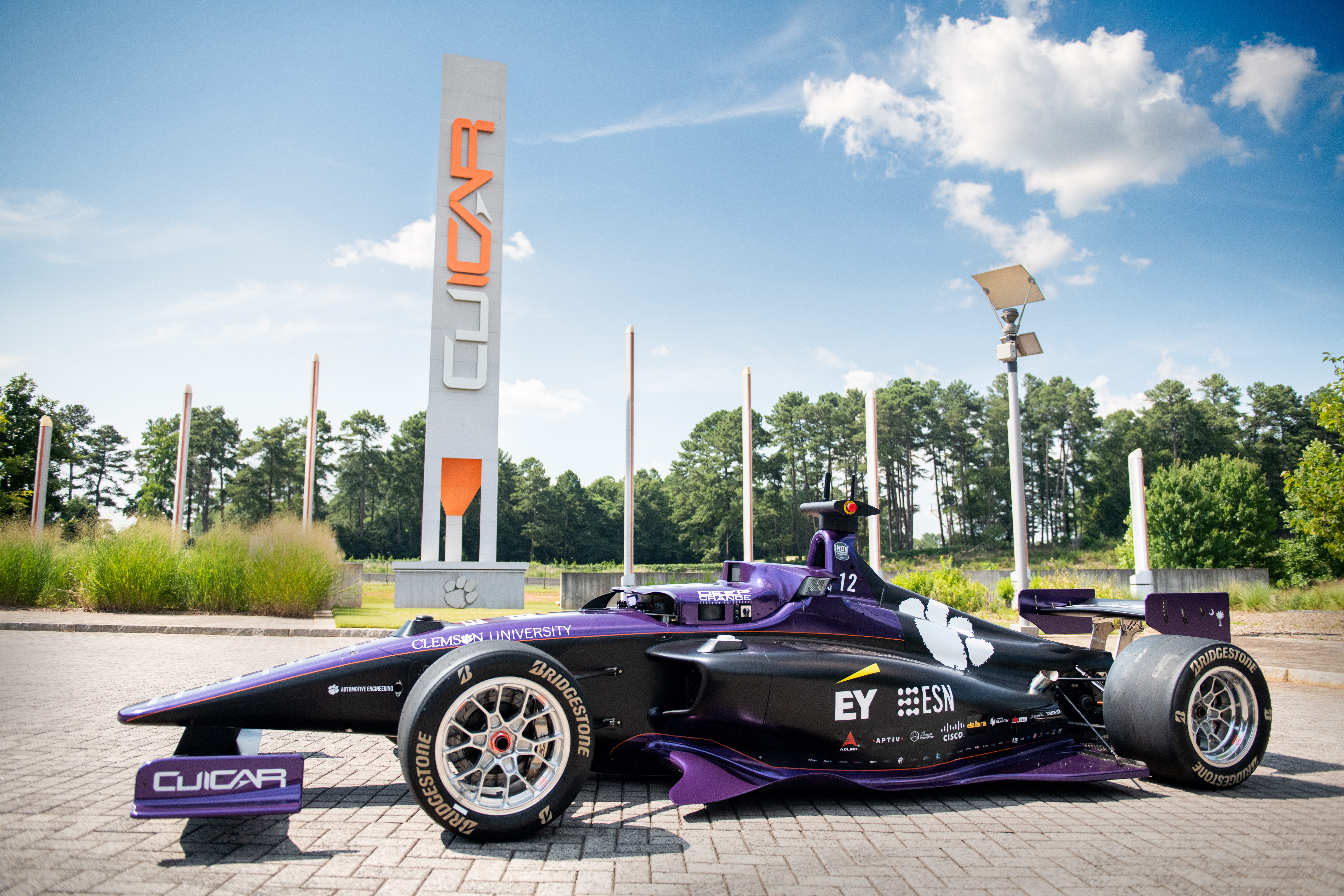 A futuristic race car that is purple and emblazoned with numerous logos,  including some that read "Clemson," "CUICAR" and "EY" is parked on an outdoor brick paver concourse with a CUICAR sign in the background. 