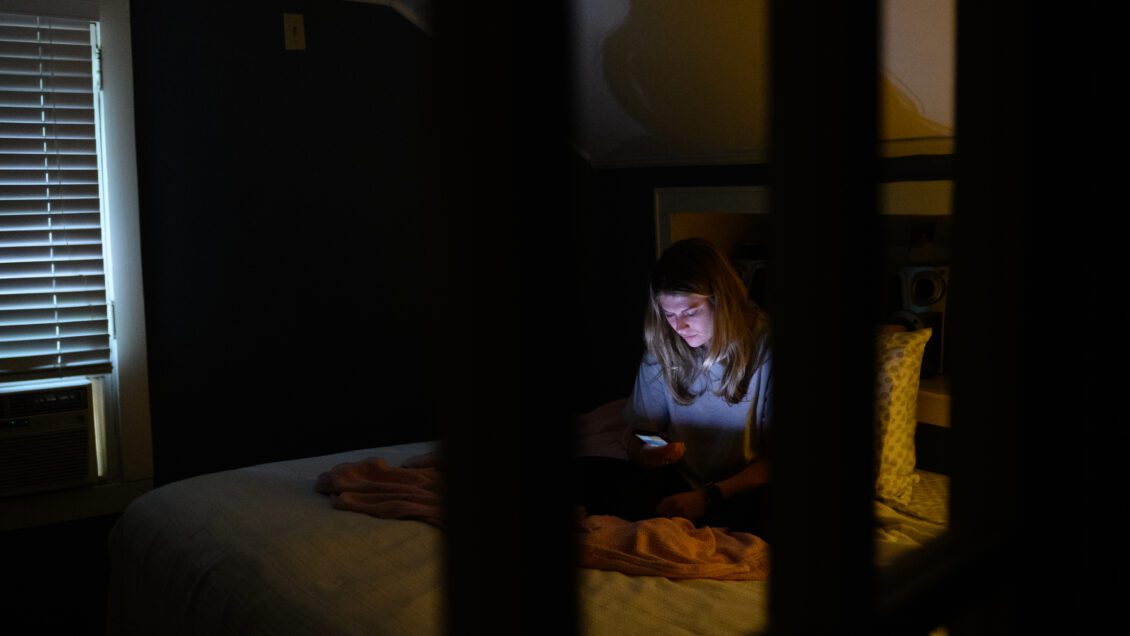 A young woman sits in a dark room on a bed looking at the lit screen of a cell phone.