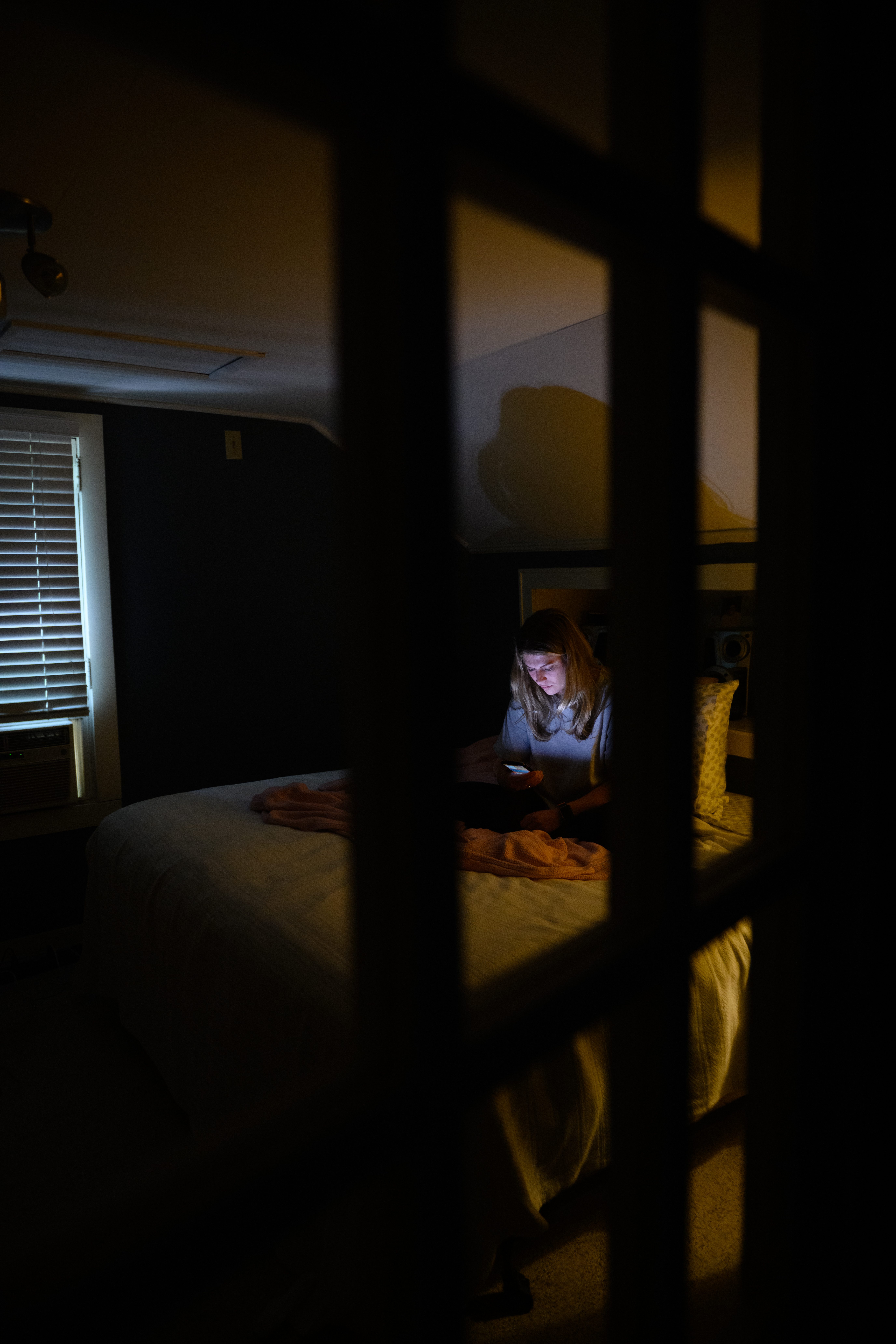 A young woman sits in a dark room on a bed looking at the lit screen of a cell phone.