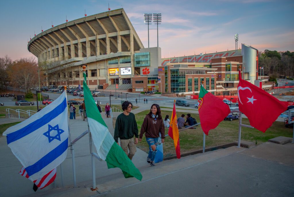 A couple walks up a staircase, flanked by rows of flags blowing in the wind, with Memorial Stadium behind them.