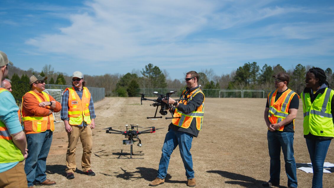 Associate professor joe Burgett holds a drone while speaking to a group of students outdoors on a sunny day.