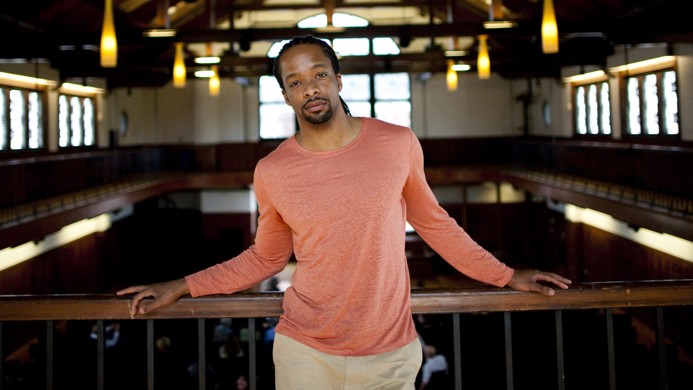 Jericho Brown stands on the balcony of a large room with vaulted ceilings and dark wooden rafters. He is a Black man with long hair pulled into a ponytail, a goatee, a salmon-colored shirt and khaki pants. His hands are resting on a railing behind him.