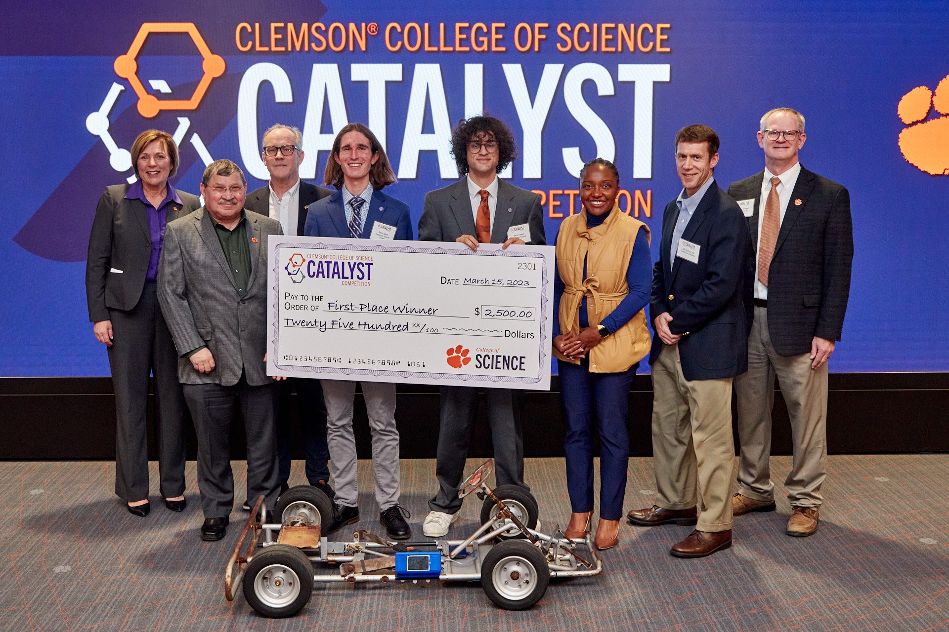 Seven people pose in front of a sign for Clemson College of Science Catalyst Competition. They are holding a big check and standing in front of a go-cart frame.