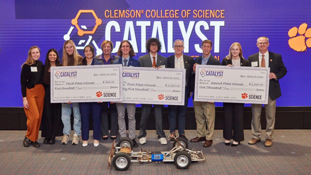 A group of people standing in front of a Catalyst Competition sign holding three big checks with a go-kart in front of them.