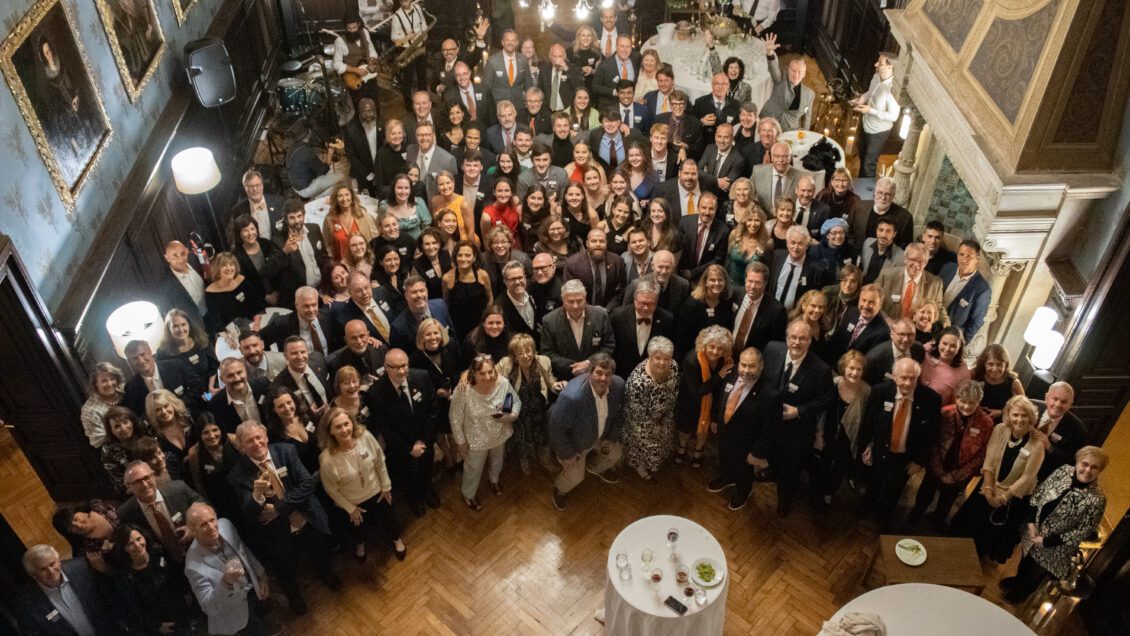 A group of more than 100 formally-dressed Clemson architecture alumni and students smile up at the camera from the floor of an ornate ballroom.