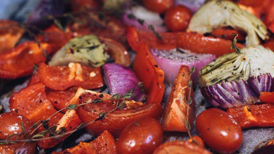 A pan of roasted vegetables including tomatoes, onions, artichokes, herbs and cracked black pepper.