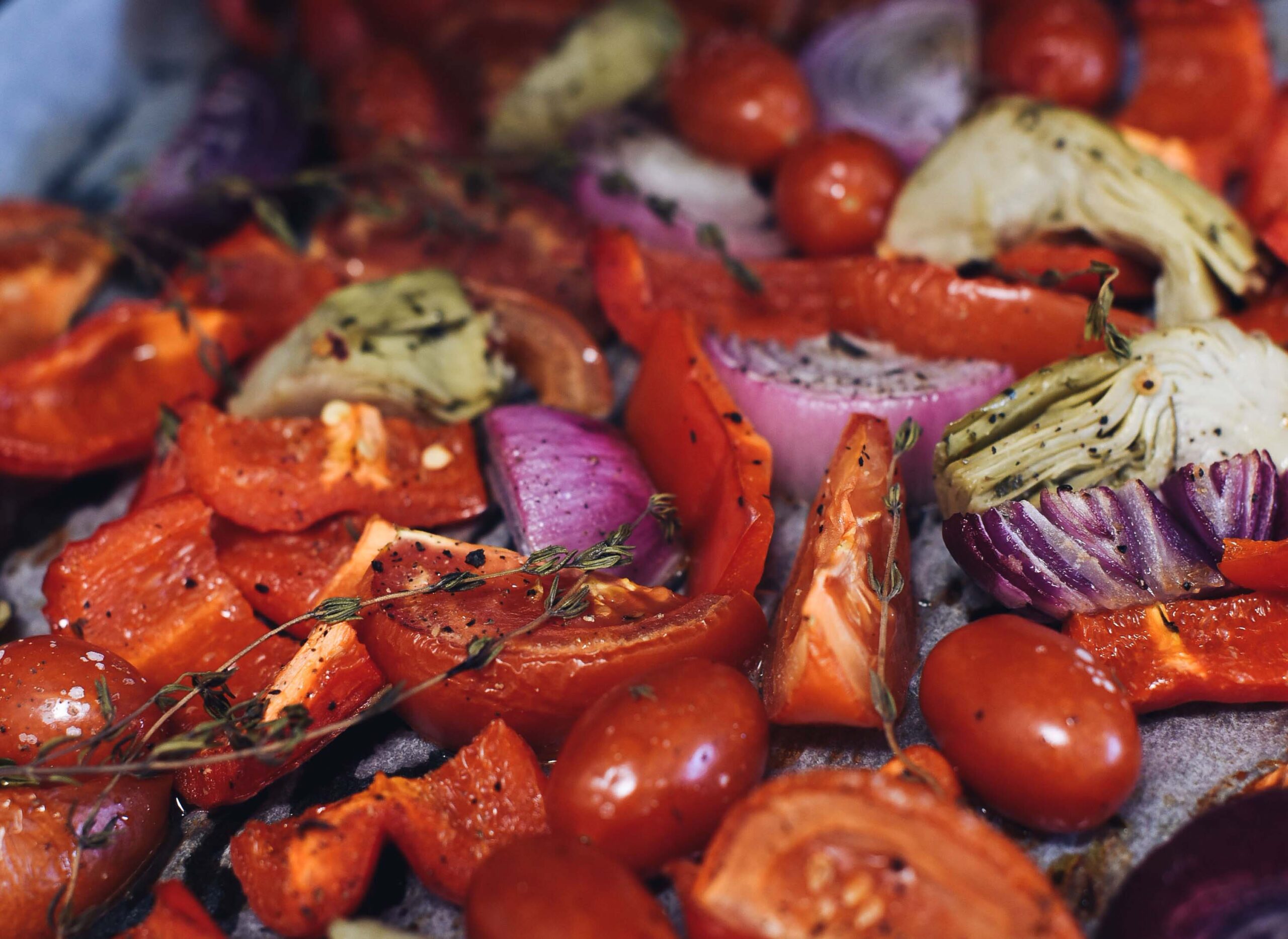 A pan of roasted vegetables including tomatoes, onions, artichokes, herbs and cracked black pepper.