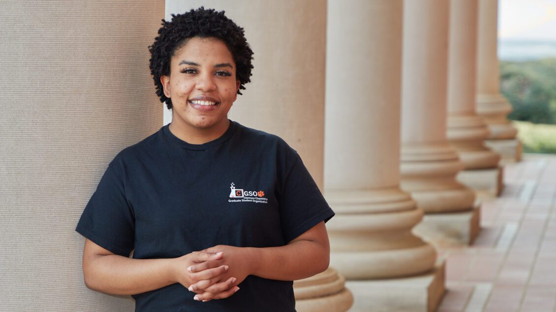 Black woman wearing a chemistry graduate student organization t-shirt leaning up against a column in front of Sikes Hall.