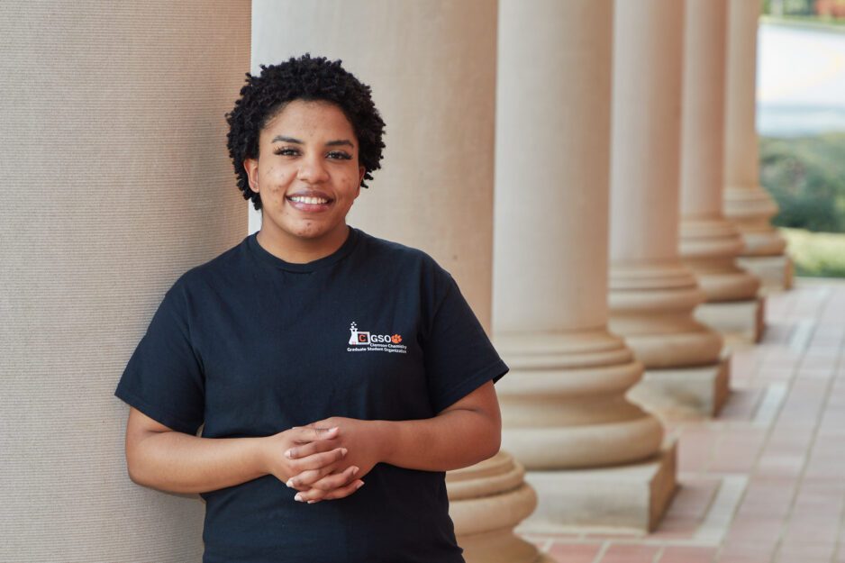 Black woman wearing a chemistry graduate student organization t-shirt leaning up against a column in front of Sikes Hall.