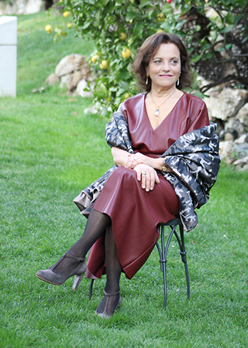 A woman with dark hair, a burgundy leather dress, and a gray floral print shawl sits on a chair in a lush green garden. She smiles and looks toward the Villa off camera.