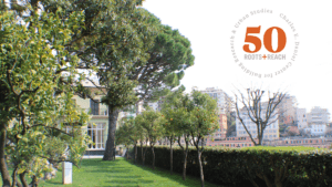 an italian villa sits behind green trees in a lush garden. The skyline of Genoa's colorful buildings can be seen over a hedge. A 50th anniversary logo is superimposed over the sky.