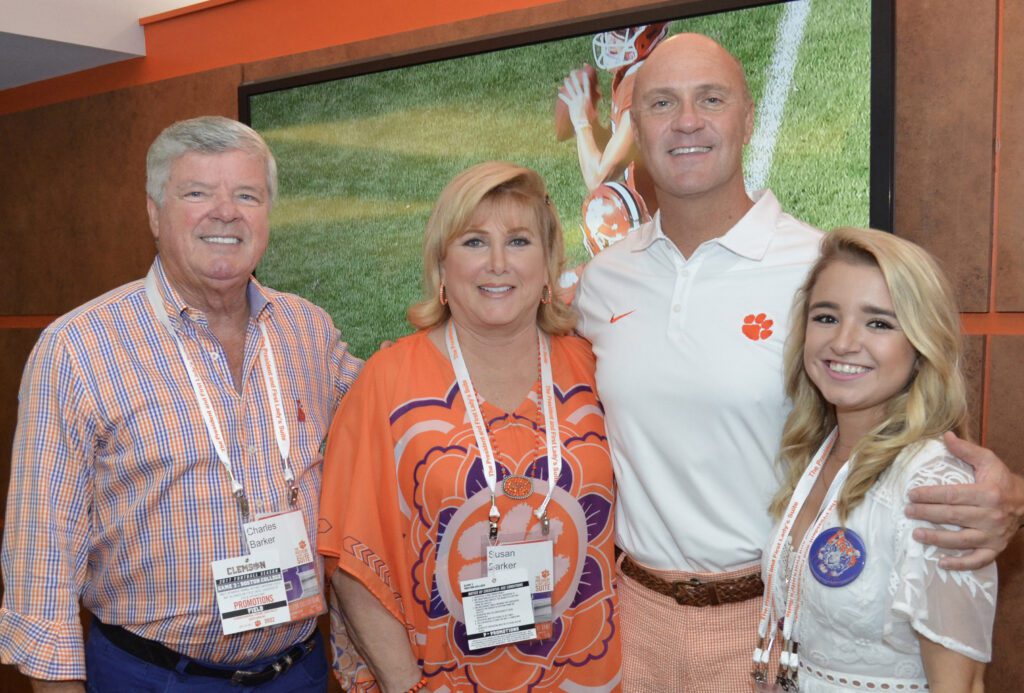 Four Caucasian people are standing inside a room in front of a large television screen with a football game on it. An older man is smiling and has his left arm across his wife's back. Standing to her right is a taller man with his right arm around her back and his left arm around a younger woman who is a Clemson alum. The two older people are her parents, and the taller man is the Clemson University President. This is the Barker Family of the Barker family Presidential Scholarship Endowment.
