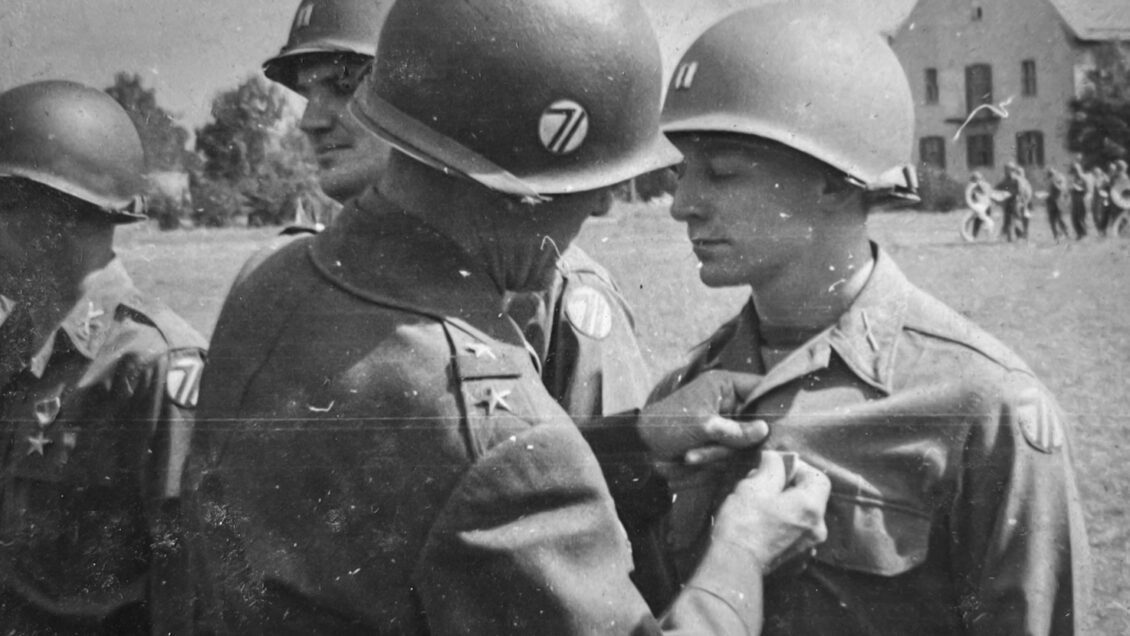 Black and white photo of a soldier pinning a Bronze Star medal on another soldier. Both wear helmets