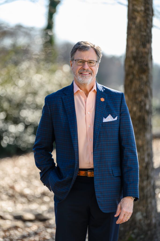 An older Caucasian man, Smith -- DSA 2022 -- is smiling. He has a short beard and mustache. His short hair is parted on the left and combed to the right. He is wearing glasses and a blazer with a pocket square. His dress shirt is unbuttoned at the top, and he is wearing a large Tiger Paw pin on his lapel. He is standing outside with a tree behind him and some bushes in the distance.