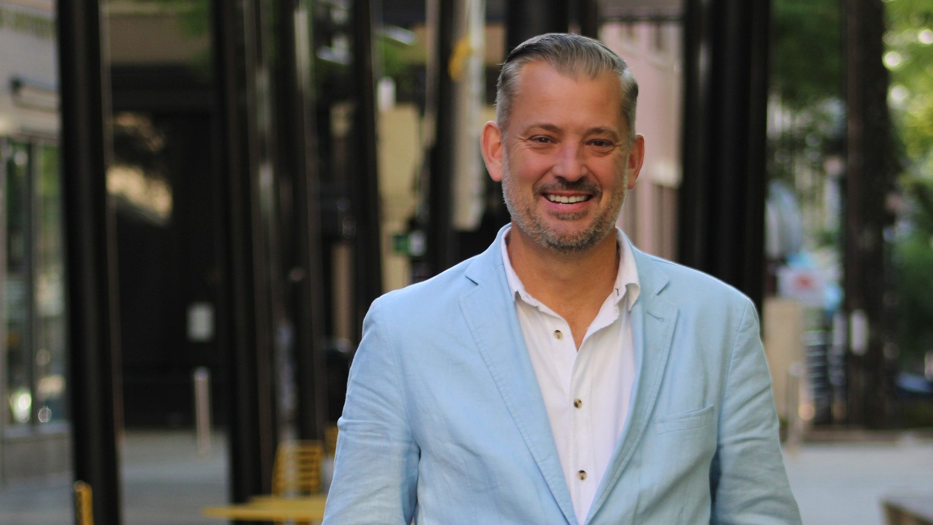 A man in a light blue blazer smiles in a city courtyard.