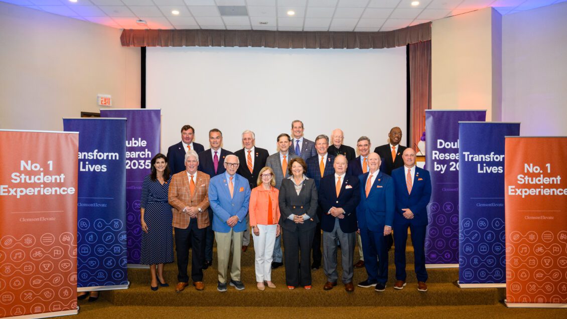A group of Clemson dignitaries including the Board of Trustees is gathered in a staging area with Clemson Elevate pop-up banners around them.