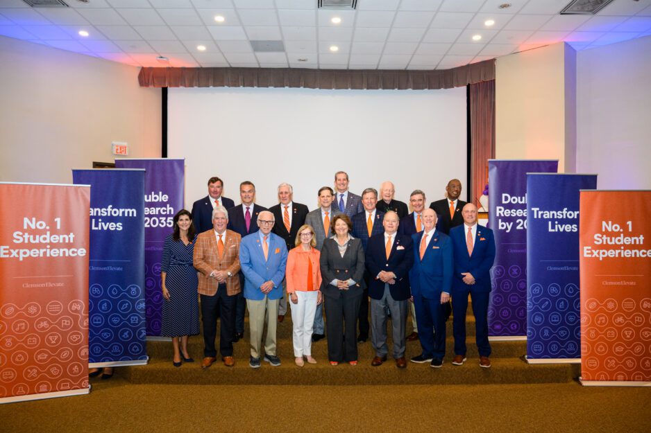 A group of Clemson dignitaries including the Board of Trustees is gathered in a staging area with Clemson Elevate pop-up banners around them.