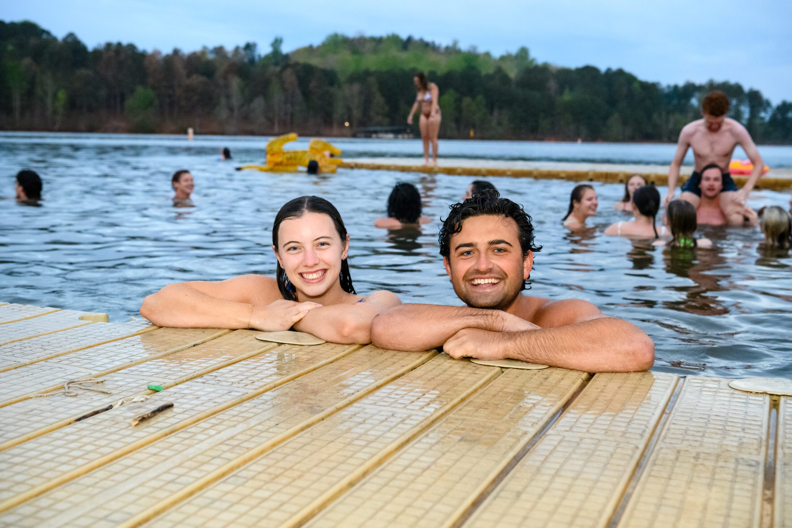A young woman and man are in the water with their arms on a dock while other students swim in the background.