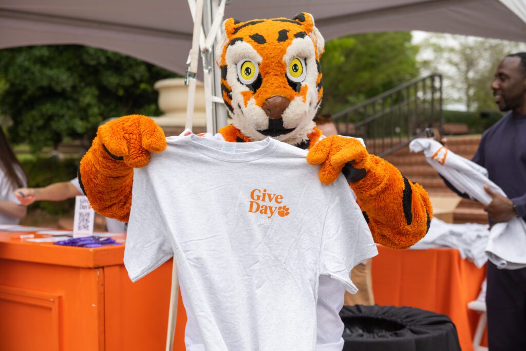 The Tiger mascot is holding a 2023 Give Day t-shirt in front of its chest. The background is a partial view of the booth and an African American man, Clemson football special team coach C.J. Spiller.