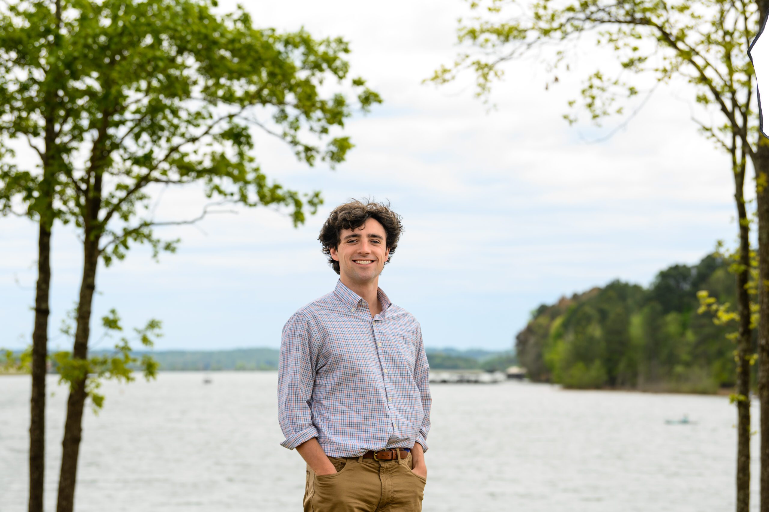 A male student wearing a light-colored dress shirt stands in front of a lake. His hands are in his pockets