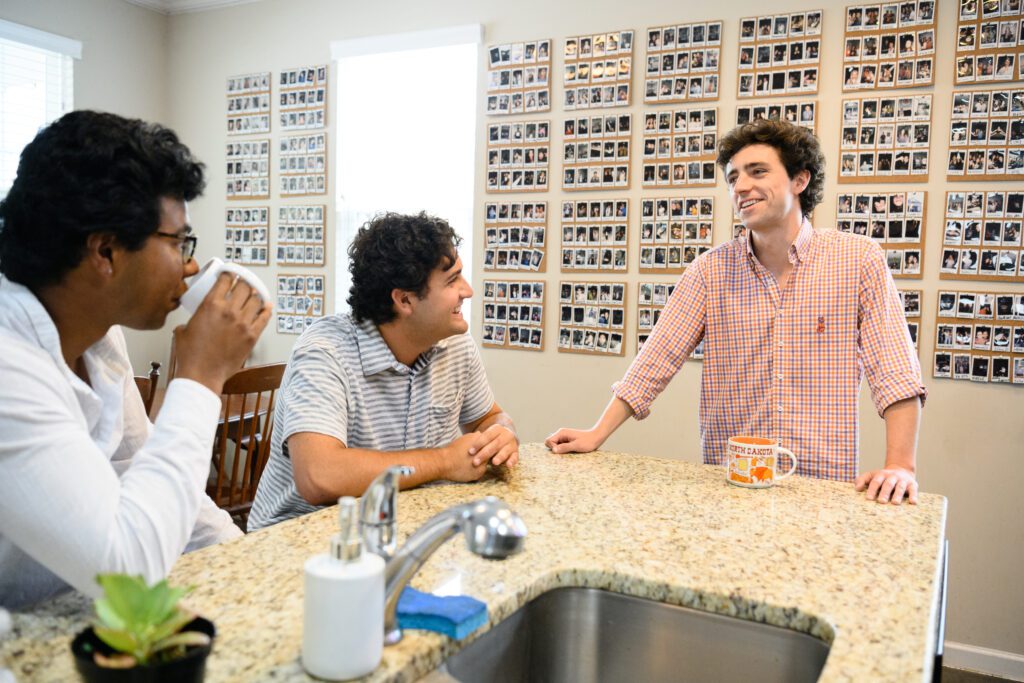 Three male college students gather around a kitchen counter with a photo wall in the background