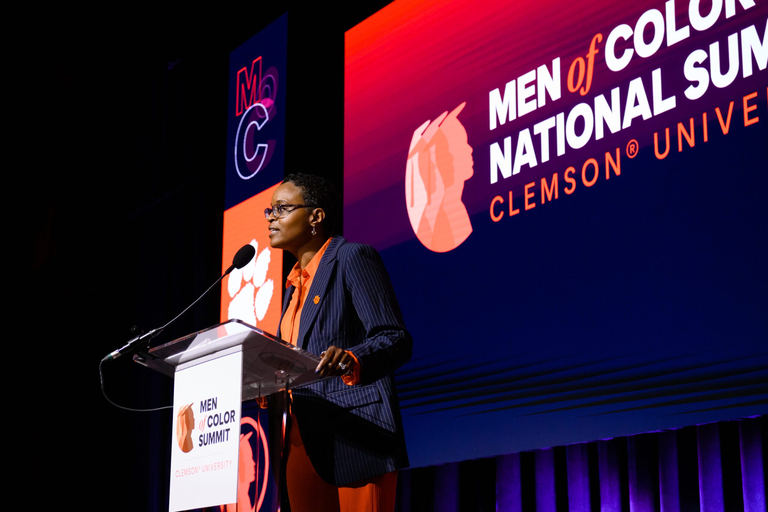 A black female stands at a podium with a Clemson University Men of Color banner hanging from it and orange and purple graphics in the background.