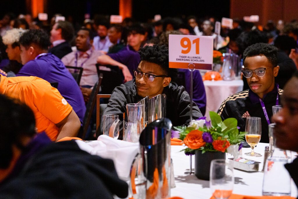 Young Black men are seated at a round table with white table cloth and a table sign with the number 91 and the words "Tiger Alliance" printed in orange. A large group of other seated individuals is seen in the background behind them. 