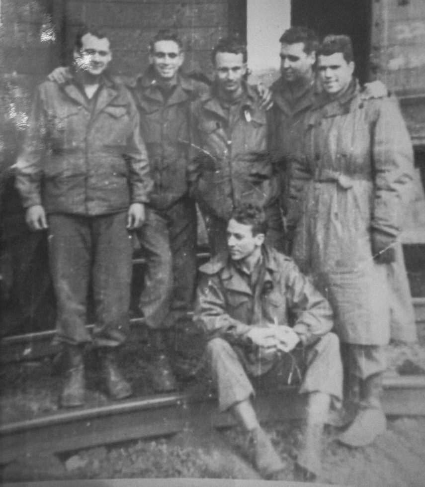 Black and white photo of six WWII soldiers in uniform standing on railroad tracks, one of them seated on a rail in front.