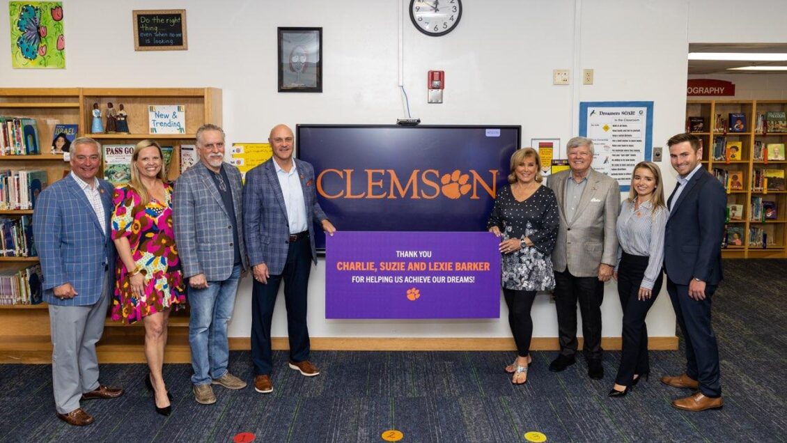 A group of eight people standing in alongside a large wall monitor/tv - four people on each side - in a school library setting. Clemson president and Susan Barker each have an end of a large poster that names the Barker Presidential Scholarship Endowment.