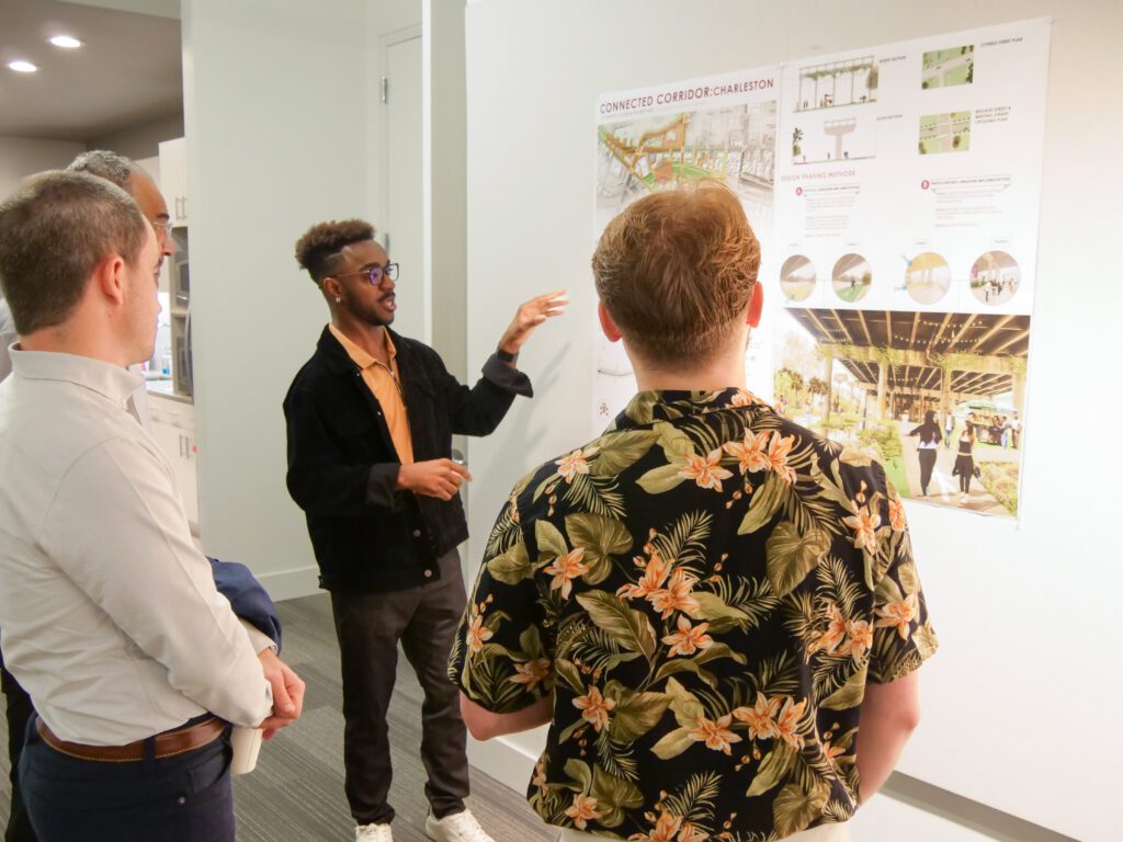 A young Black man with glasses gestures toward a poster showing images of an urban park. Three men--two white and one Black--stand and listen to him speak. They are in a large white room with accent lighting. 