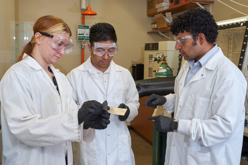 Three men wearing white lab coats and black gloves look at something in a lab at the Clemson Nanomaterials Institute.