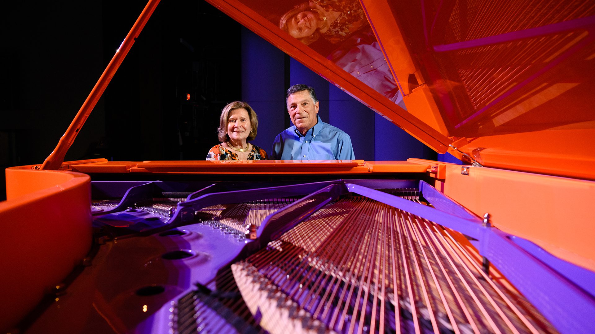 A distinguished couple--man and woman--smile while seated at an orange and purple piano. The interior of the piano is purple, and the exterior is orange. The photo looks under the lid of the piano to show the strings in the foreground.