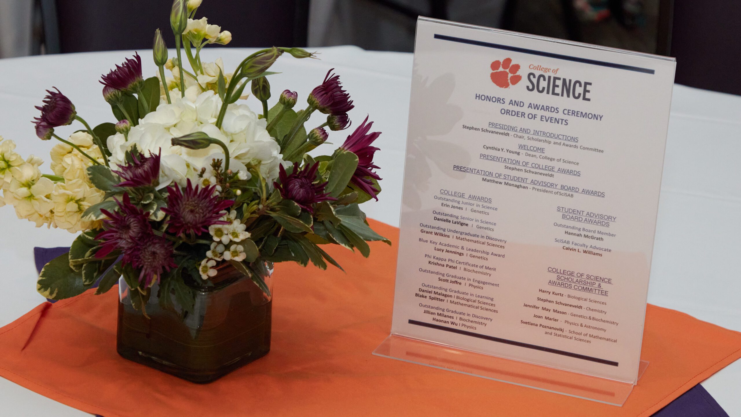 A photograph of a white flower table setting with a piece of paper in an acrylic holder showing the order of the program at the College of Science's student Honors and Awards ceremony.