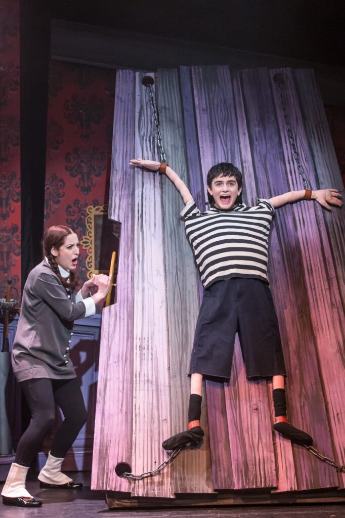 A stage shows a boy wearing a black and white striped shirt strapped with chains to a wooden wall while a girl with pigtails appears to grab a handle attached to the wall. 