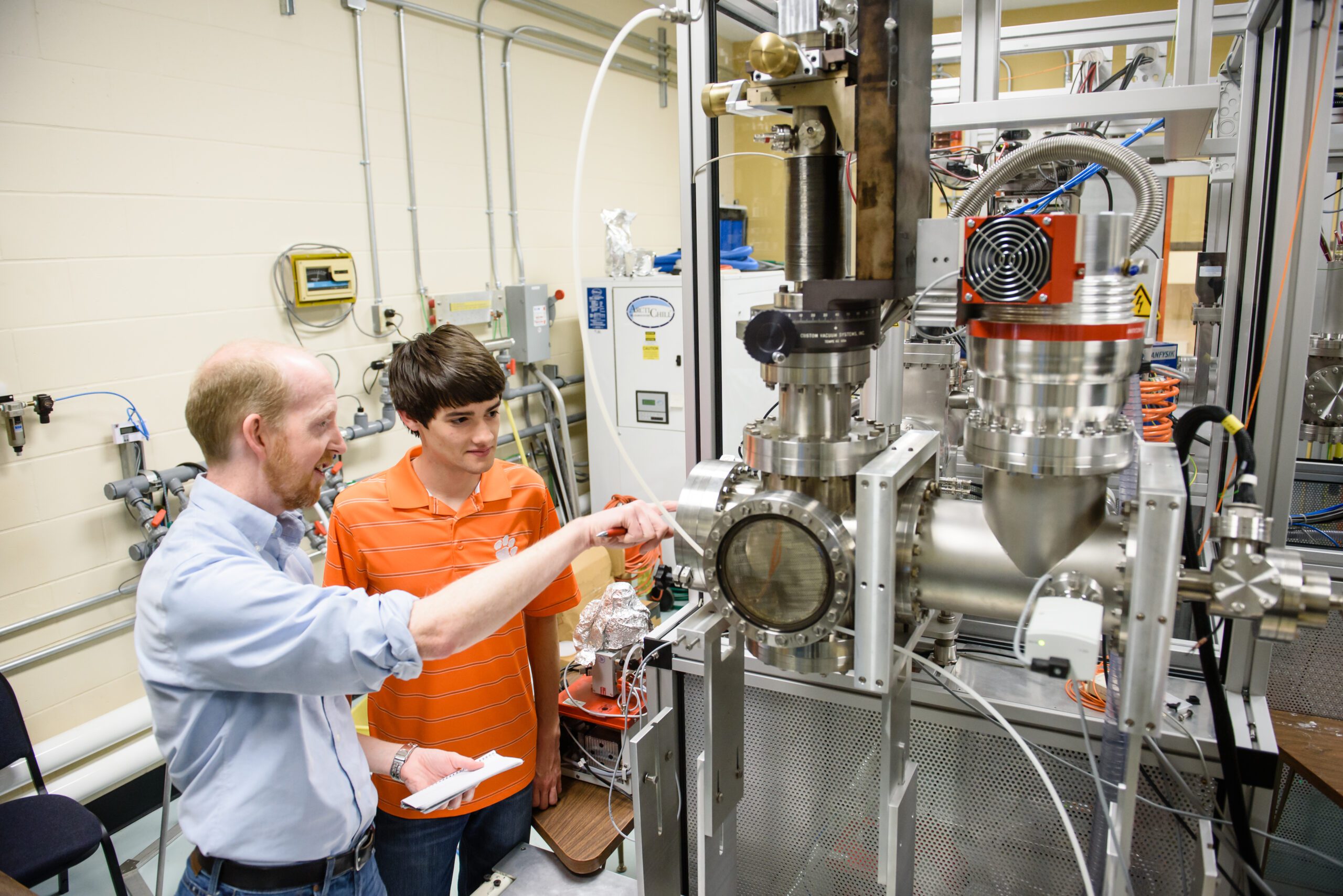 A male faculty wearing a blue dress shirt rolled up at the sleeves works with a male student wearing an orange polo bearing a Tiger paw, reading a temperature gauge on a large piece of scientific equipment. 