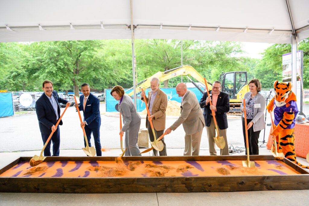 Men and women wearing  dress clothes are lined up and using shovels for a ceremonial groundbreaking into an orange raised box. 