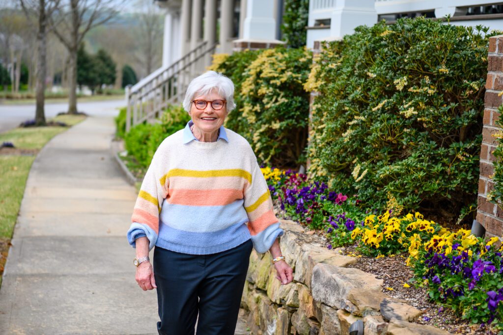 A woman with white hair wearing glasses and a pastel sweater walks along a stone wall with yellow flowers spilling over it. 
