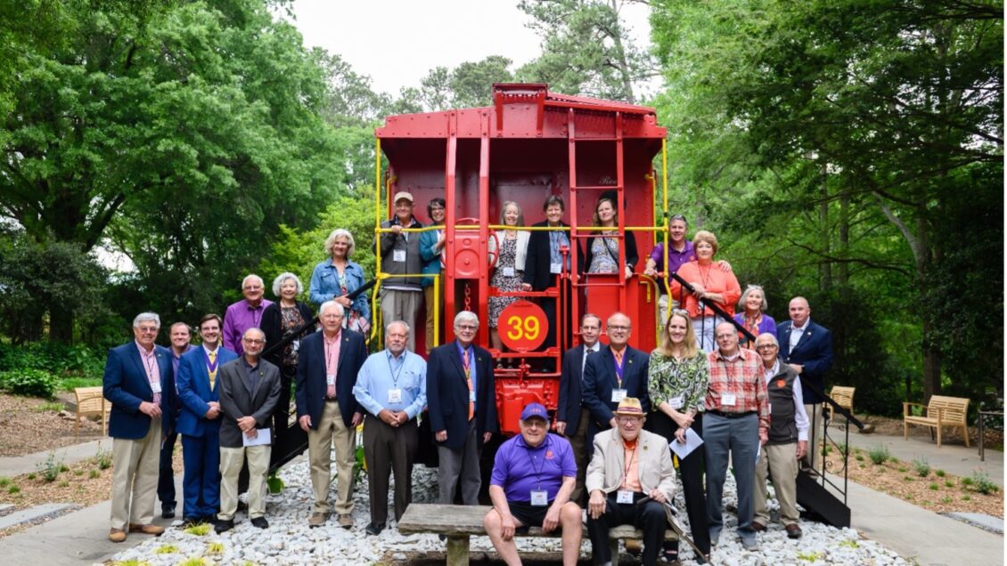 A group casually stands smiling for a photographer around the front of an old, red train that is no longer in use but that is now part of a garden landscape as an attraction for area residents.