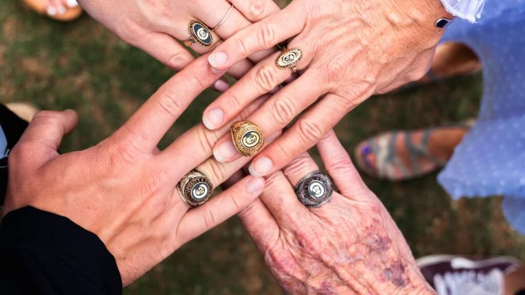 Four hands reach into the frame from each corner wearing Clemson rings, one with two rings