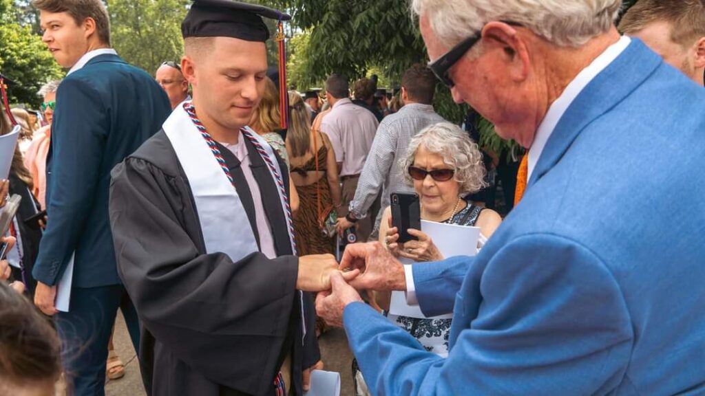 A man in a suit turns the ring on the finger of a man in graduation gowns and cap.