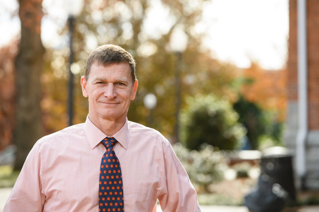 An environmental portrait of a white male standing on campus, wearing an orange dress shirt and tiger paw print tie